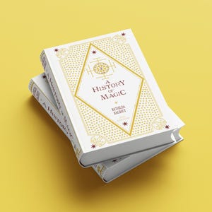 Puede incluir: Dos libros de tapa dura apilados titulados A History of Magic por Bathilda Bagshot, 2nd Edition. Las cubiertas blancas presentan patrones geométricos dorados, un marco central en forma de diamante y pequeñas estrellas rojas. El título aparece en los lomos.
