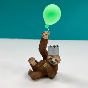 May include: A brown sloth figurine wearing a silver party hat and holding a green balloon.
