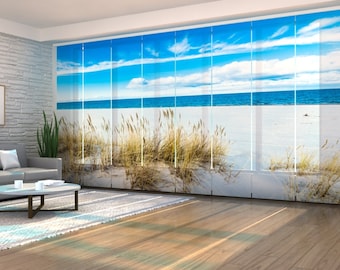 May include: Vertical panel blinds displaying a beach scene. The image shows a bright blue sky with white clouds above a calm, blue sea. The foreground features a sandy beach with tall, dry grasses, creating a serene coastal view.