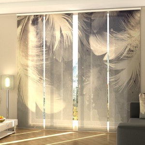 May include: White and gray patterned window shades with a feather design. The shades are hanging in a living room with a couch and a lamp.