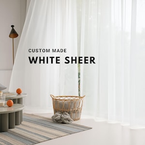 May include: White sheer curtains hang in front of a window, with the text "CUSTOM MADE WHITE SHEER" in the upper left. A woven basket with a gray lining and two gray yarn balls sit on a striped rug. Two small round tables with oranges are in the foreground.