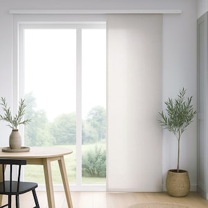 Op de afbeelding: Een lichte, luchtige interieurscène met een groot raam en een wit gordijn. Een ronde houten tafel met een zwarte stoel staat op de voorgrond, en een potplant voegt groen toe. De kamer is gevuld met natuurlijk licht.