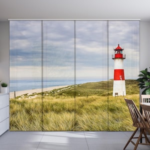 May include: A red and white lighthouse stands tall on a sandy beach with green grass and a blue ocean in the background.