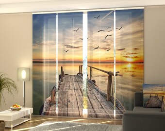 May include: A set of sheer window curtains featuring a printed image of a wooden jetty stretching out over a tranquil body of water at sunset. The sky is a vibrant orange and yellow, with birds flying overhead.