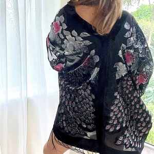 May include: A black sheer kimono with a floral print and fringe trim. The kimono has a white and pink floral design with a peacock feather pattern.