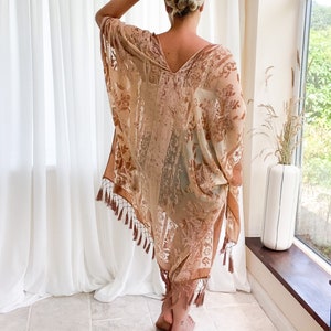 May include: A sheer, flowing, beige kimono with a floral pattern and a V-neck. The kimono has brown tassels along the hem and sleeves. The garment is worn over other clothing. The background includes white curtains and a window.