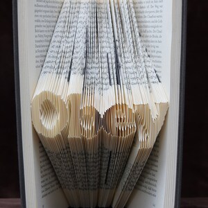 May include: A book with pages folded into the shape of the word "LOVE". The book is closed and standing upright on a brown surface.