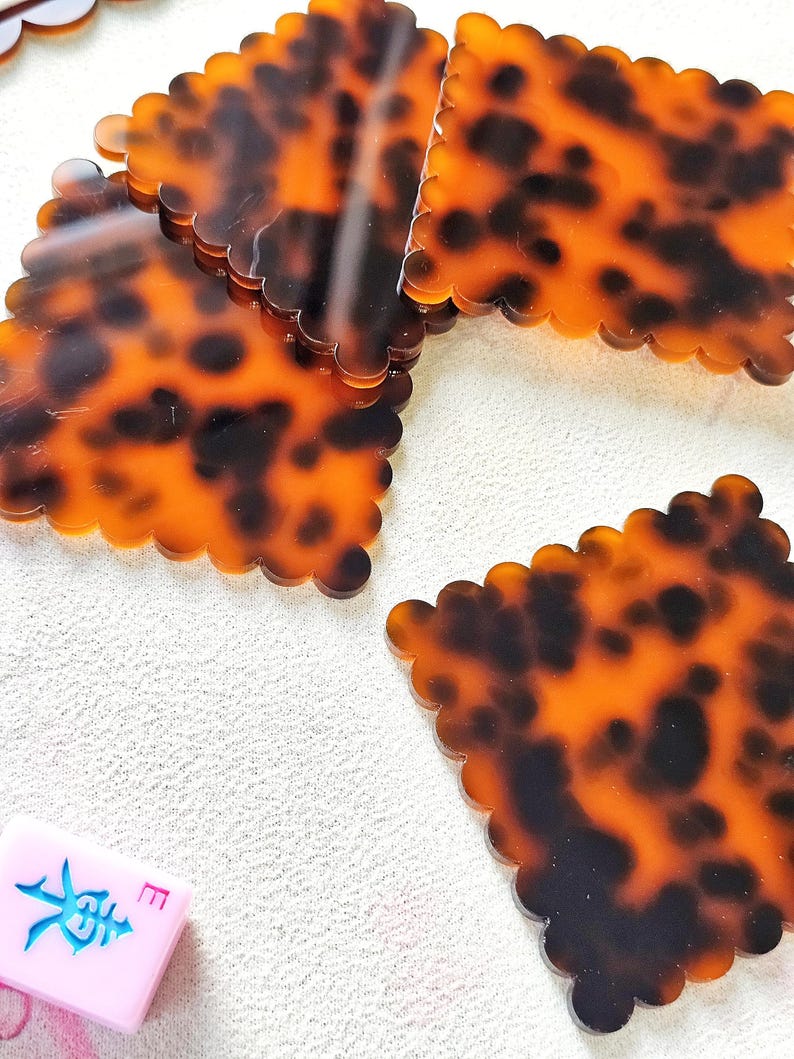 May include: Several tortoise shell patterned coasters with scalloped edges are scattered on a white surface. A pink Mahjong tile with blue Chinese characters is also visible. The coasters have a glossy finish.