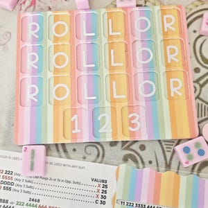 May include: A pastel-coloured Mahjong set with a rainbow striped design. The game board spells out "ROLLOR" and displays the numbers "1 2 3". Includes pink tiles and a paper with game rules. A fun and colourful game.