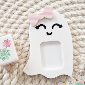 May include: A white ghost-shaped cutout with a smiling face and a pink and white striped bow attached to the top. Next to the ghost is a rectangular white tile featuring the number two in blue and two floral designs in red and green ink.