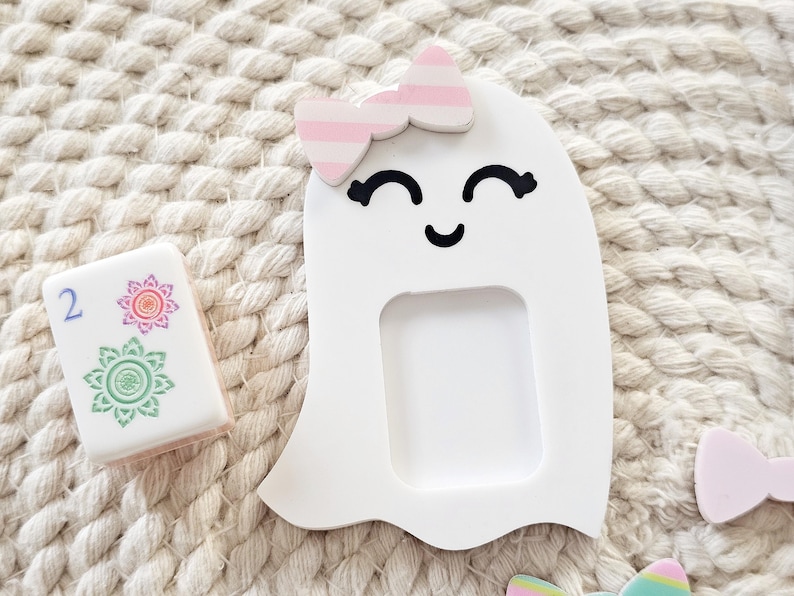May include: A white ghost-shaped cutout with a smiling face and a pink and white striped bow attached to the top. Next to the ghost is a rectangular white tile featuring the number two in blue and two floral designs in red and green ink.