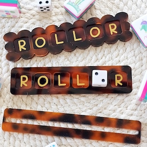 Puede incluir: Una colecci&oacute;n de piezas de juego, que incluyen dados, fichas y dos tableros de juego "ROLLOR" con un patr&oacute;n de carey. La palabra "ROLLOR" est&aacute; escrita en letras doradas en los tableros. Las piezas del juego est&aacute;n dispuestas sobre una superficie texturizada de color blanquecino.