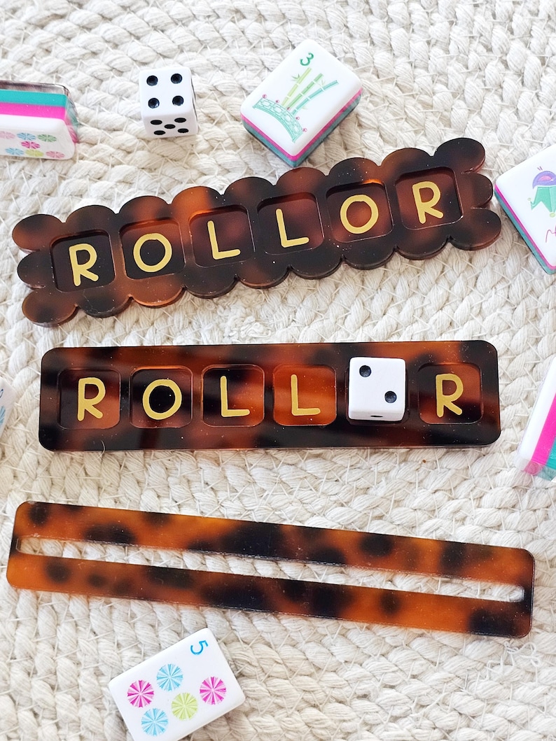 Puede incluir: Una colecci&oacute;n de piezas de juego, que incluyen dados, fichas y dos tableros de juego "ROLLOR" con un patr&oacute;n de carey. La palabra "ROLLOR" est&aacute; escrita en letras doradas en los tableros. Las piezas del juego est&aacute;n dispuestas sobre una superficie texturizada de color blanquecino.