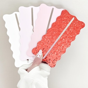 May include: A hand holds a fan of decorative paper bookmarks. The bookmarks are in shades of white, pink, and red. The red bookmark has a glittery texture. Each bookmark has a wavy edge and a vertical slit.