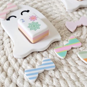 May include: A white ghost-shaped cutout with a pink and white striped bow on its head and a smiling face. A rectangular tile with the number 2 and two floral designs sits on the ghost. Several small, colorful bows with stripes and solid colors are scattered nearby.