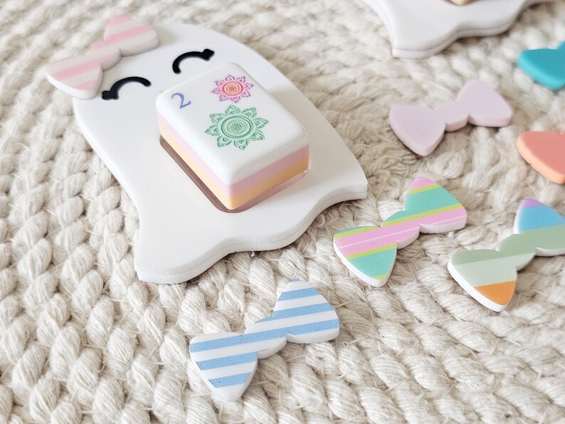 May include: A white ghost-shaped cutout with a pink and white striped bow on its head and a smiling face. A rectangular tile with the number 2 and two floral designs sits on the ghost. Several small, colorful bows with stripes and solid colors are scattered nearby.