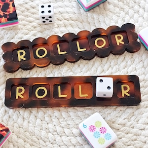 Puede incluir: Primer plano de un juego de mesa con dados, fichas y dos bandejas marrones "ROLLOR". Las bandejas tienen un patr&oacute;n de carey con letras doradas. Un dado blanco con puntos negros y otras fichas coloridas son visibles.