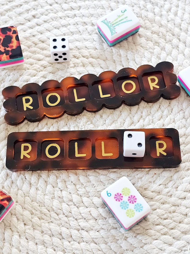 Puede incluir: Primer plano de un juego de mesa con dados, fichas y dos bandejas marrones "ROLLOR". Las bandejas tienen un patr&oacute;n de carey con letras doradas. Un dado blanco con puntos negros y otras fichas coloridas son visibles.