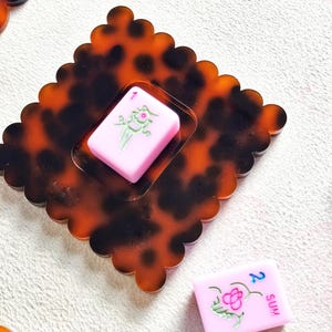 May include: A decorative, tortoise-shell-patterned tray with a scalloped edge, holding a pink Mahjong tile with a green floral design. Another pink tile with the number 2 and the word "SUM" is also visible.