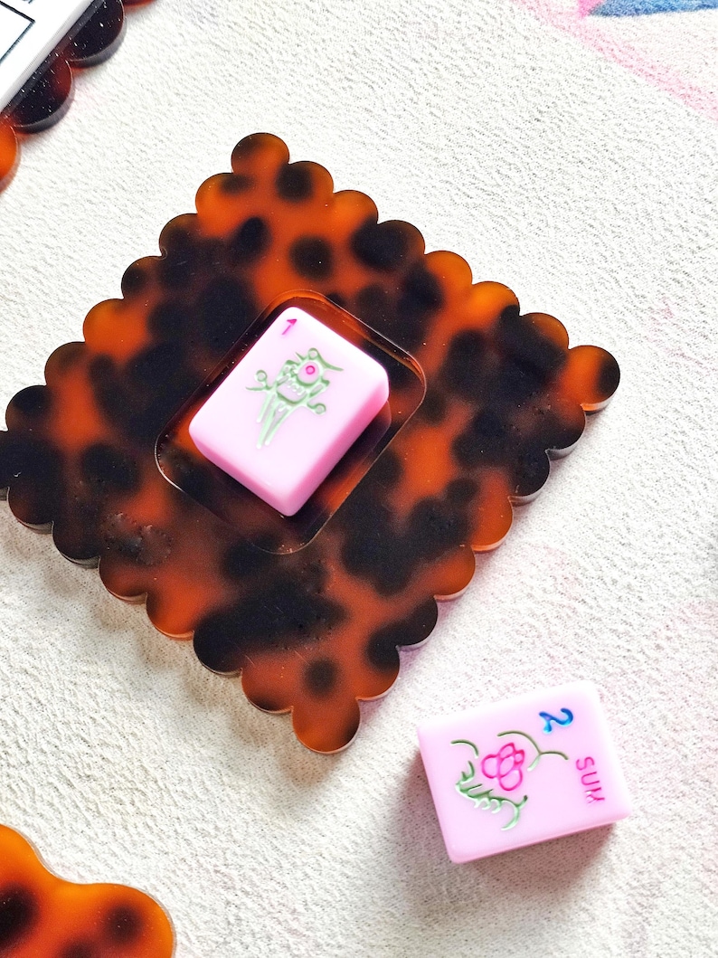 May include: A decorative, tortoise-shell-patterned tray with a scalloped edge, holding a pink Mahjong tile with a green floral design. Another pink tile with the number 2 and the word "SUM" is also visible.