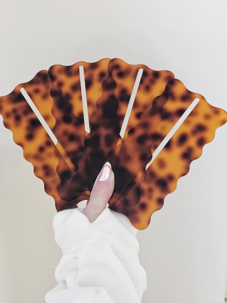 Peut inclure: Une main tient cinq pinces &agrave; cheveux en &eacute;caille de tortue. Les pinces ont une forme d'&eacute;ventail avec un bord festonn&eacute; et des barres verticales blanches. Les pinces sont marron et ambre avec des taches sombres.