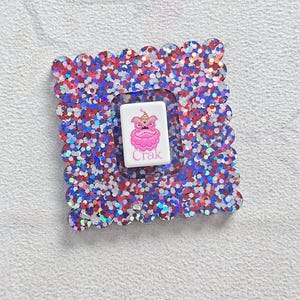 Peut inclure: Un cadre carr&eacute; et scintillant avec un bord festonn&eacute;, rempli de paillettes rouges, blanches et bleues. &Agrave; l'int&eacute;rieur, un rectangle blanc pr&eacute;sente une cr&eacute;ature rose de dessin anim&eacute; avec une couronne et le mot "Crak".
