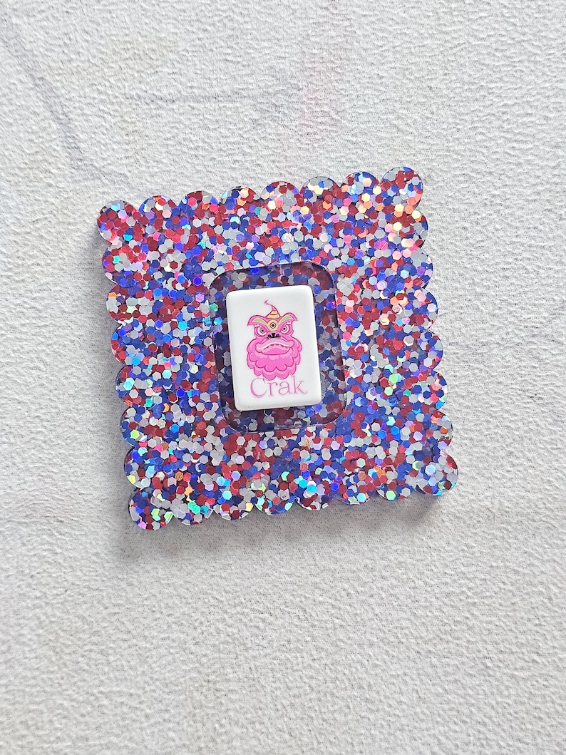 Peut inclure: Un cadre carr&eacute; et scintillant avec un bord festonn&eacute;, rempli de paillettes rouges, blanches et bleues. &Agrave; l'int&eacute;rieur, un rectangle blanc pr&eacute;sente une cr&eacute;ature rose de dessin anim&eacute; avec une couronne et le mot "Crak".