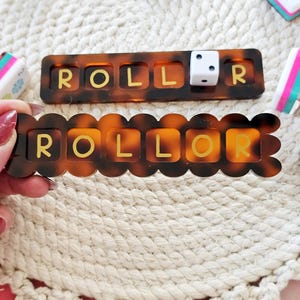 Puede incluir: Dos piezas de juego color carey con la palabra "ROLLOR" en letras doradas. Una pieza es rectangular y la otra tiene un borde festoneado. Un dado blanco est&aacute; en la pieza rectangular. Otros dados y gomas de borrar est&aacute;n en el fondo.