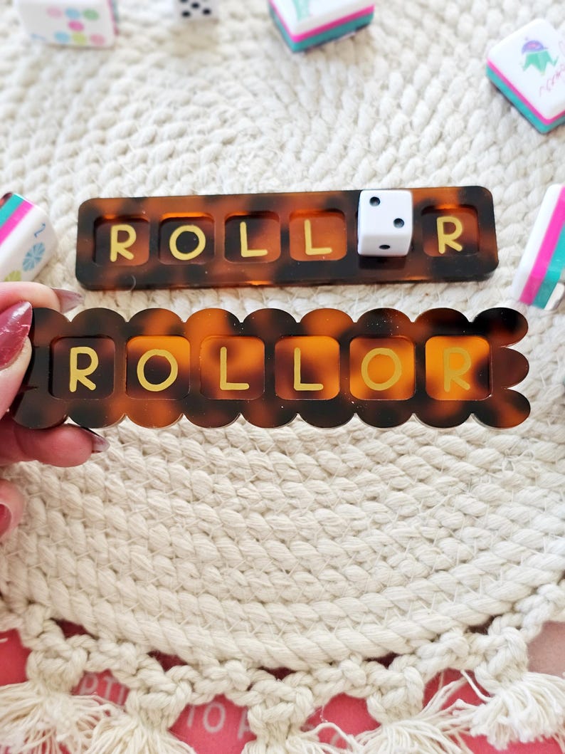 Puede incluir: Dos piezas de juego color carey con la palabra "ROLLOR" en letras doradas. Una pieza es rectangular y la otra tiene un borde festoneado. Un dado blanco est&aacute; en la pieza rectangular. Otros dados y gomas de borrar est&aacute;n en el fondo.