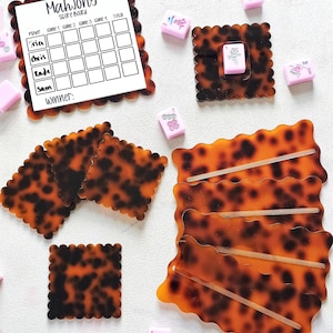 May include: A Mahjong game set featuring a score board, tiles, and holders. The pieces have a tortoiseshell design. The score board is white with the word "Mahjong" at the top. Pink tiles are scattered around the board.