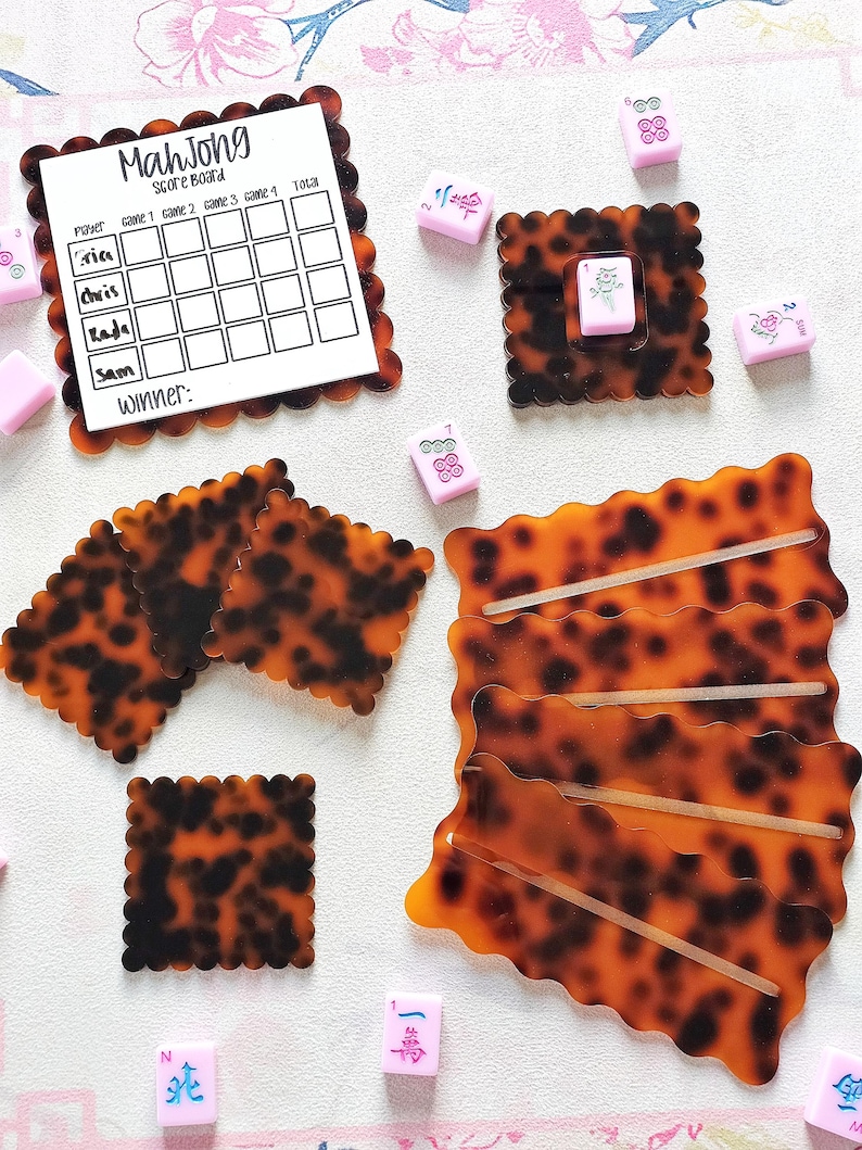 May include: A collection of Mahjong game pieces and accessories. The set includes a score board with the word "Mahjong" at the top, along with several tiles and holders in a tortoiseshell pattern. Pink tiles are scattered around the board.
