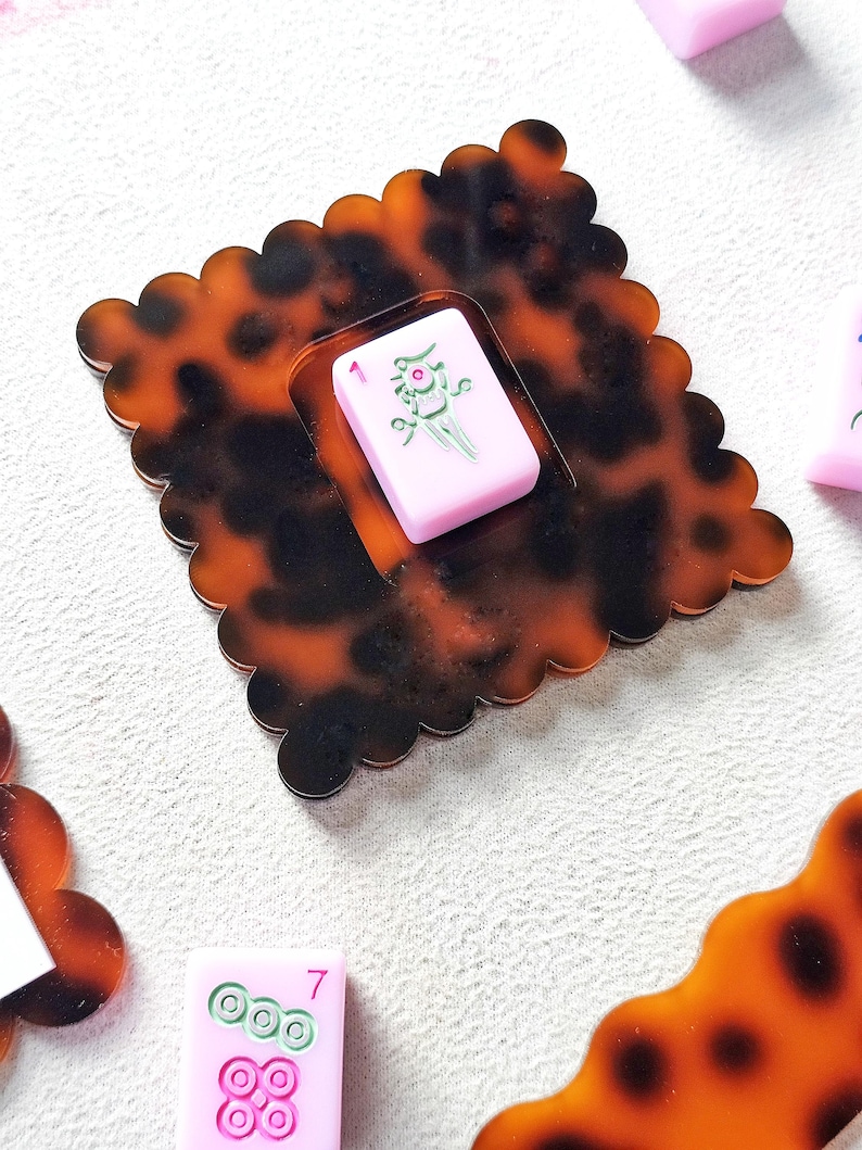 May include: A decorative, tortoise-shell-patterned coaster with a scalloped edge. A pink Mahjong tile with a green floral design and the number 1 is set in the center. Additional pink Mahjong tiles are visible.
