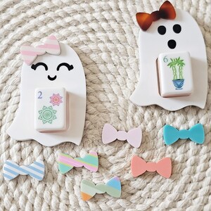 May include: Two white ghost-shaped figures with black facial features, each holding a mahjong tile. One ghost wears a pink striped bow, the other a brown tortoiseshell bow. Six additional small, colorful bows in various patterns are arranged below.