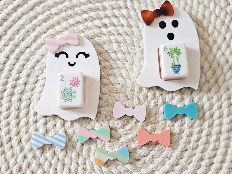 May include: Two white ghost-shaped figures with black facial features, each holding a mahjong tile. One ghost wears a pink striped bow, the other a brown tortoiseshell bow. Six additional small, colorful bows in various patterns are arranged below.
