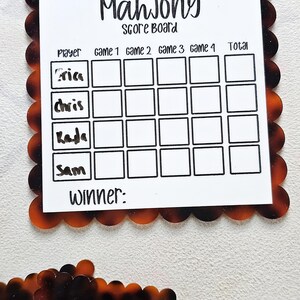 May include: A Mahjong score board with a white center and a decorative tortoiseshell border. The board has spaces for players' names and game scores, with the word "Mahjong" at the top. The names "Aria", "Chris", "Kada", and "Sam" are listed.