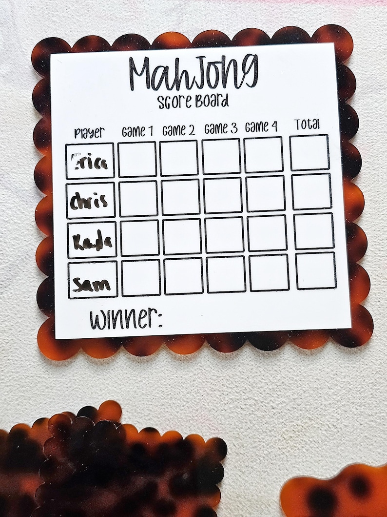 May include: A Mahjong score board with a white center and a decorative tortoiseshell border. The board has spaces for players' names and game scores, with the word "Mahjong" at the top. The names "Aria", "Chris", "Kada", and "Sam" are listed.