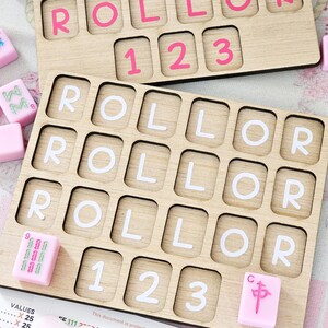 May include: Two rectangular wooden boards with grid cutouts for tiles. The top board features pink letters and numbers, while the bottom board displays white letters and numbers. Several loose pink tiles with green and pink symbols are scattered nearby.
