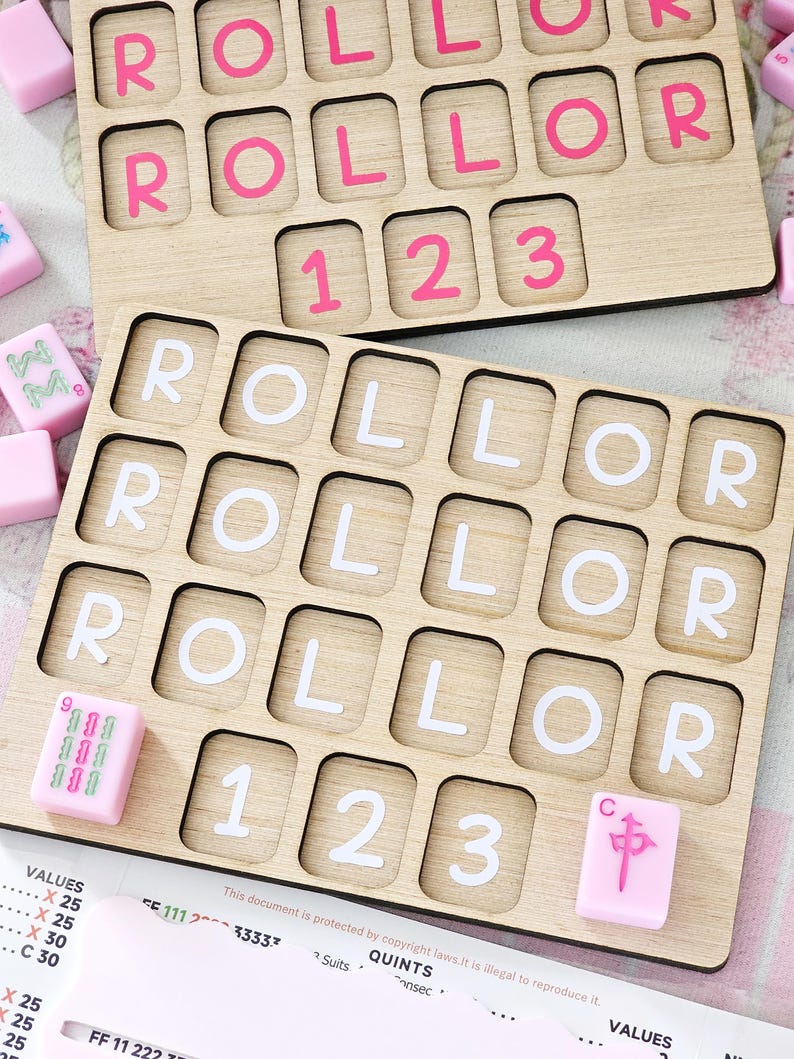 May include: Two rectangular wooden boards with grid cutouts for tiles. The top board features pink letters and numbers, while the bottom board displays white letters and numbers. Several loose pink tiles with green and pink symbols are scattered nearby.