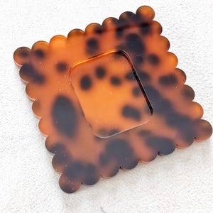 Puede incluir: Un posavasos cuadrado con estampado de carey y borde festoneado. El posavasos tiene un recorte cuadrado transparente en el centro. La paleta de colores incluye tonos marrones y negros, creando un accesorio decorativo &uacute;nico.