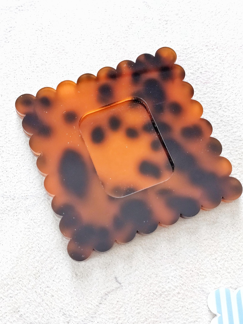 Puede incluir: Un posavasos cuadrado con estampado de carey y borde festoneado. El posavasos tiene un recorte cuadrado transparente en el centro. La paleta de colores incluye tonos marrones y negros, creando un accesorio decorativo &uacute;nico.