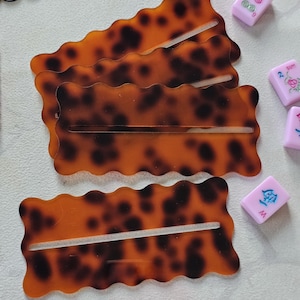 May include: Four tortoise shell-patterned hair clips with a wavy edge and a slot in the center. Several pink Mahjong tiles with various symbols are scattered around the clips. The clips are a warm brown color with dark brown spots.