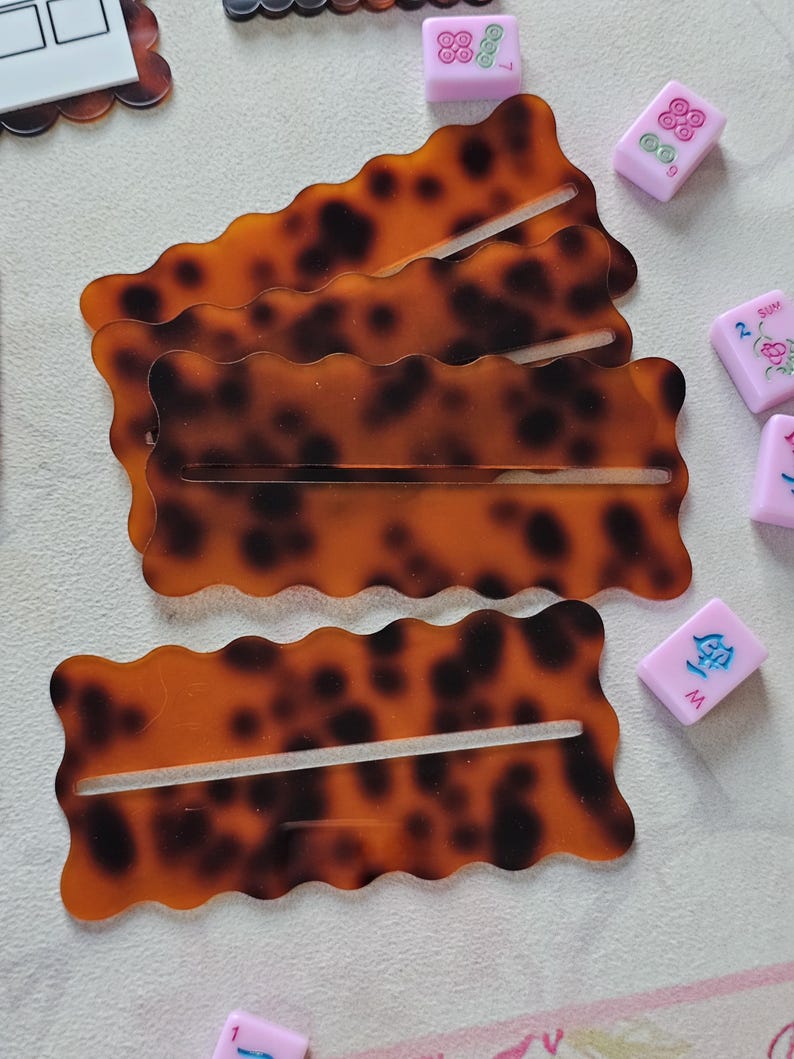 May include: Four tortoise shell-patterned hair clips with a wavy edge and a slot in the center. Several pink Mahjong tiles with various symbols are scattered around the clips. The clips are a warm brown color with dark brown spots.