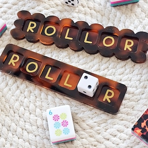 Puede incluir: Dos piezas de juego color carey con la palabra "ROLLOR" en letras doradas. Tambi&eacute;n hay dos dados blancos y dos fichas de juego. Las piezas y fichas del juego est&aacute;n sobre una superficie tejida blanca.