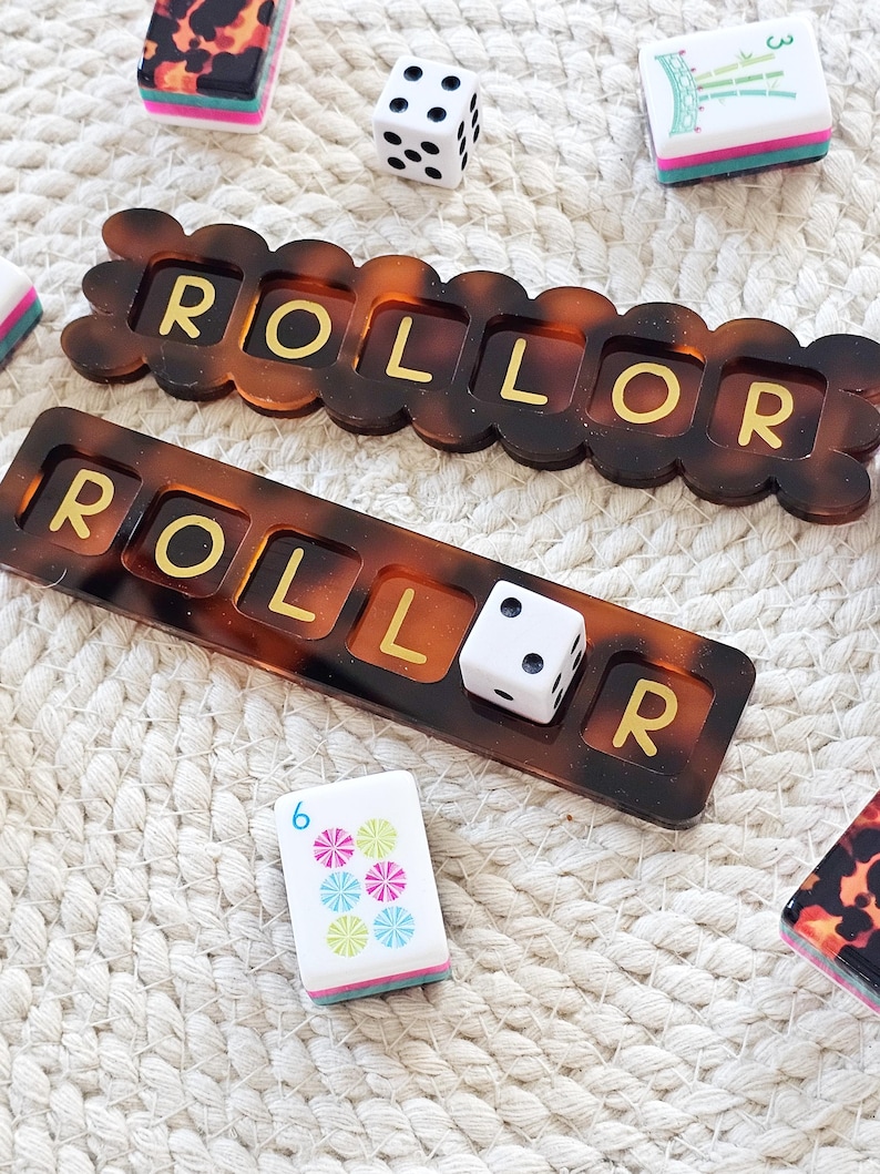 Puede incluir: Dos piezas de juego color carey con la palabra "ROLLOR" en letras doradas. Tambi&eacute;n hay dos dados blancos y dos fichas de juego. Las piezas y fichas del juego est&aacute;n sobre una superficie tejida blanca.