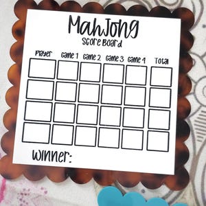 May include: A Mahjong score board with a brown tortoiseshell scalloped border. The white center has the words "Mah Jong Score Board" and boxes to record player scores for each game. The word "Winner:" is at the bottom.