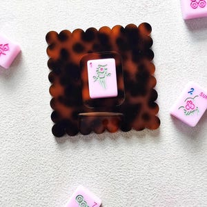 May include: A tortoiseshell-patterned hair clip with a scalloped edge, holding a pink Mahjong tile with a green and white design. Scattered around are other pink Mahjong tiles with various symbols and numbers. A fun accessory for game lovers.