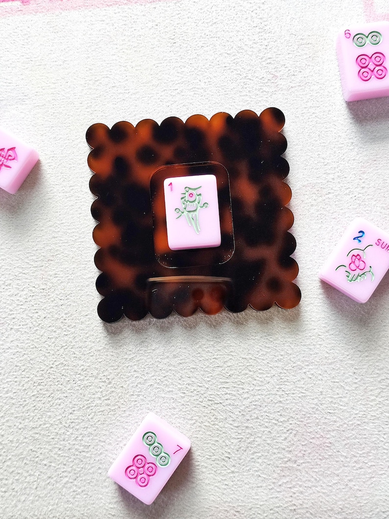 May include: A tortoiseshell-patterned hair clip with a scalloped edge, holding a pink Mahjong tile with a green and white design. Scattered around are other pink Mahjong tiles with various symbols and numbers. A fun accessory for game lovers.