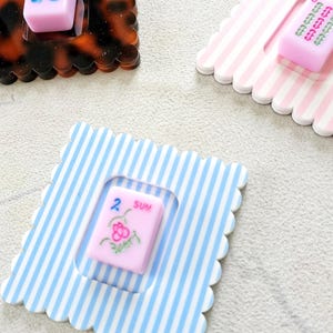 Puede incluir: Un conjunto de posavasos decorativos con fichas de Mahjong. Los posavasos tienen bordes festoneados y presentan varios patrones, incluyendo carey, rayas rosas y blancas, y rayas azules y blancas. Las fichas de Mahjong son rosas con varios s&iacute;mbolos.