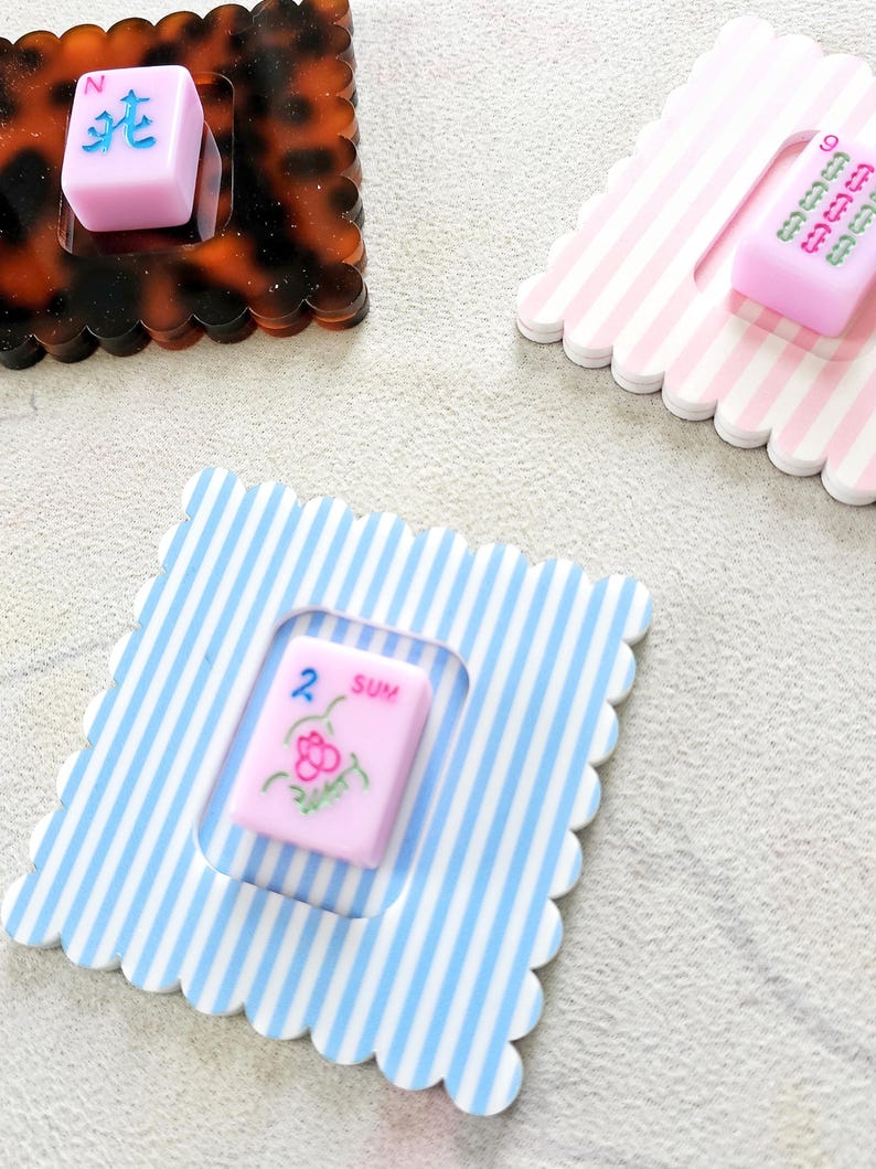 Puede incluir: Un conjunto de posavasos decorativos con fichas de Mahjong. Los posavasos tienen bordes festoneados y presentan varios patrones, incluyendo carey, rayas rosas y blancas, y rayas azules y blancas. Las fichas de Mahjong son rosas con varios s&iacute;mbolos.