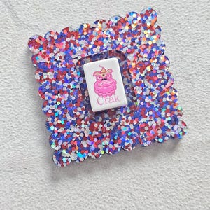 May include: A square coaster with a scalloped edge, filled with red, white, and blue glitter. The coaster has a white, rectangular insert in the centre with a pink cartoon character and the word "Crak".